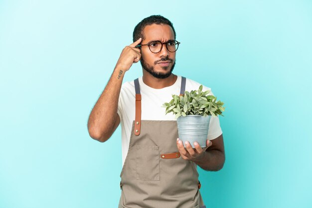 Hombre latino jardinero sosteniendo una planta aislada de fondo azul con dudas y pensando