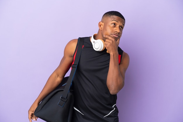 Hombre latino joven deporte con bolsa de deporte aislado en la pared púrpura que tiene dudas