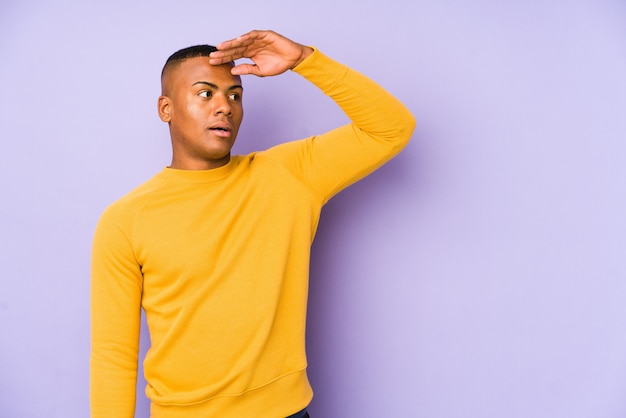 Hombre latino joven en la pared púrpura que mira lejos manteniendo la mano en la frente.