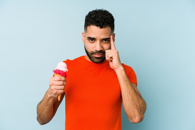 Hombre latino joven que sostiene un helado que señala el templo con el dedo, pensando, centrado en una tarea.