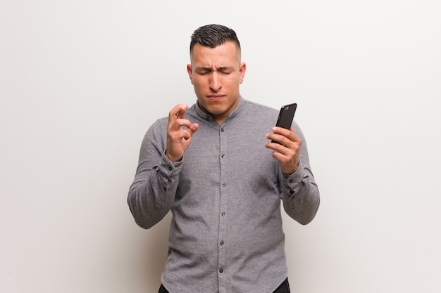 Hombre latino joven que sostiene un teléfono que cruza los dedos para tener suerte