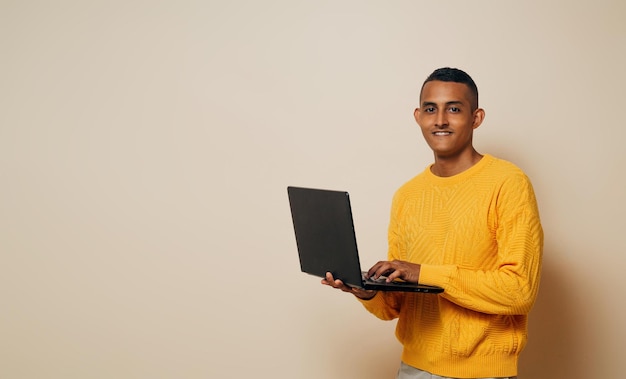 Hombre latino joven sosteniendo una computadora portátil mirando a la cámara sobre un fondo marrón con espacio de copia