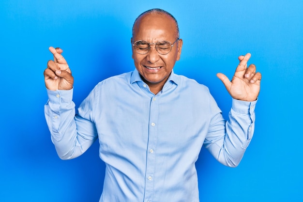 Hombre latino de mediana edad con ropa casual y gafas gestando con el dedo cruzado sonriendo con esperanza y ojos cerrados suerte y concepto supersticioso