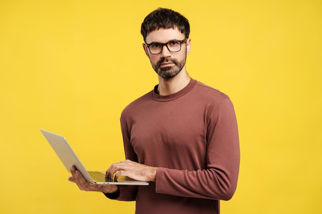 Hombre latino pensativo especialista en IT con gafas sosteniendo una computadora portátil aislada sobre un fondo amarillo
