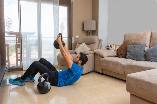 Hombre latino que realiza un entrenamiento de cross fit en casa con un par de pesas rusas.