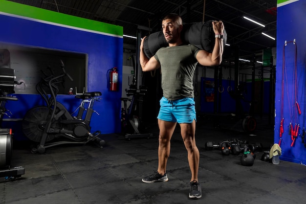 Hombre latino con ropa deportiva que lleva un saco de arena en la espalda mientras entrena en un gimnasio