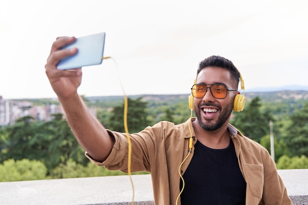 Foto hombre latino con teléfono celular en la mano haciendo una videollamada hombre latino con barba y gafas de sol con auriculares amarillos