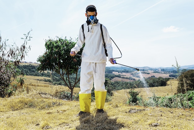 Hombre latino vistiendo ropa adecuada y máscara protectora rociando malezas en el terreno