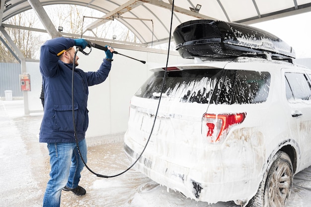 Hombre lavando agua a alta presión coche todoterreno americano con portaequipajes en lavado de autoservicio en clima frío