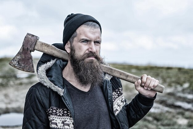 Hombre leñador. Hipster hombre guapo o chico con barba y bigote en rostro serio con sombrero y chaqueta sostiene hacha oxidada con empuñadura de madera al aire libre en la cima de la montaña contra el cielo nublado sobre fondo natural