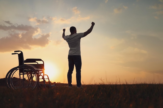 Hombre levantando las manos hacia el cielo cerca de la silla de ruedas al atardecer vista trasera Milagro curativo