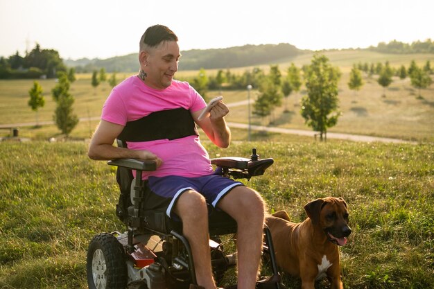 Hombre LGBT con discapacidad usando un teléfono inteligente mientras su perro yace cerca de él en el parque al atardecer