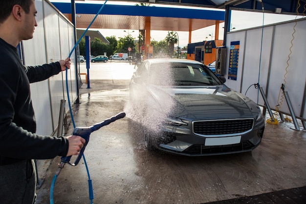 Hombre limpiando automóviles de lujo con agua a alta presión en una gasolinera de lavado al aire libre en el fondo y hombre con pistola de agua en primer plano