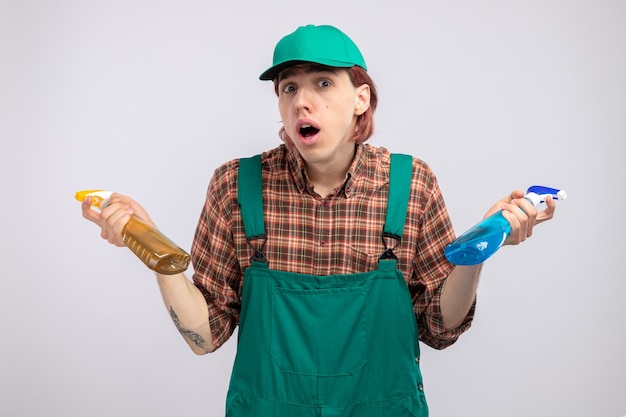 Hombre de limpieza joven en mono de camisa a cuadros y gorra sosteniendo botellas de spray de limpieza confundido teniendo dudas encogiéndose de hombros de pie sobre la pared blanca