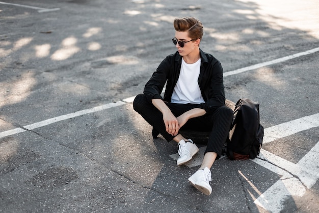 Hombre lindo joven inconformista en ropa elegante elegante negra con gafas de sol de moda