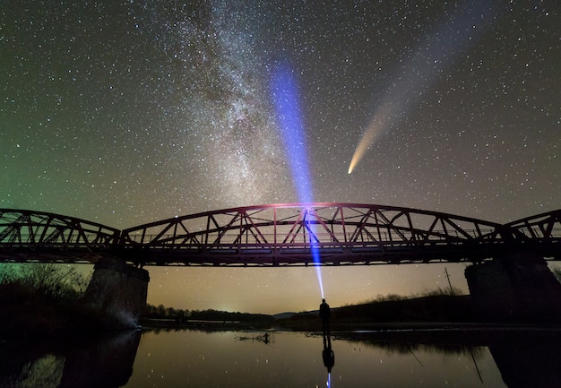 Hombre con linterna de pie en la orilla del río bajo el puente de metal iluminado bajo el oscuro cielo estrellado y el cometa Neowise con cola de luz reflejada en el agua.