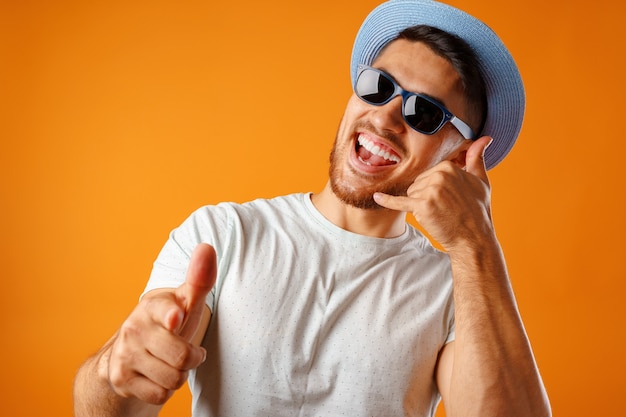 Hombre lleno de alegría con sombrero de paja y gafas de sol mostrando gesto de llamarme contra el fondo amarillo