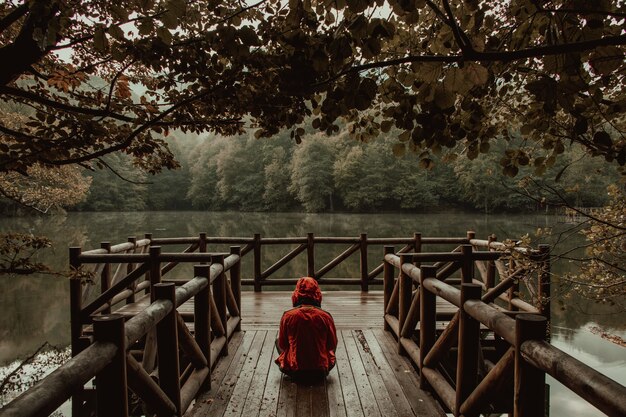 El hombre lleva chaqueta roja y está sentado en un puente de madera junto al lago