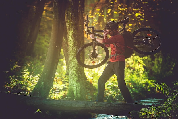 Foto hombre llevando una bicicleta en el tronco de un árbol sobre un arroyo en el bosque