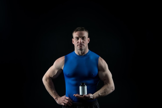 Hombre macho guapo o chico musculoso sexy con cuerpo de atleta y torso en chaleco deportivo azul sostiene tarro de pastillas para vitamina esteroide drud o dopaje sobre fondo negro