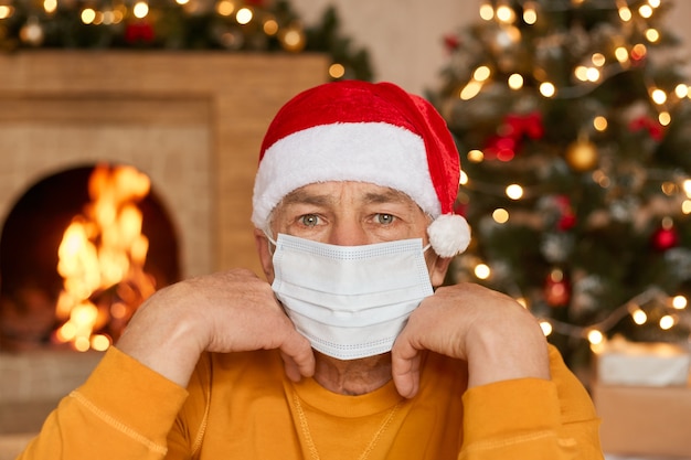 Hombre maduro aburrido sentado en la sala de estar con decoración de Año Nuevo, manteniendo los puños debajo de la barbilla, con máscara de seguridad médica para la prevención de infecciones, camisa amarilla y gorro de santa.