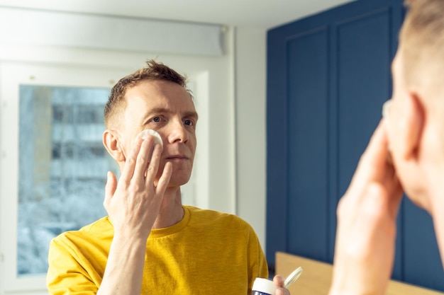 Hombre maduro, aplique tónico facial con una almohadilla de algodón y luego masajee con las manos Retrato masculino en el espejo de la habitación Limpieza facial