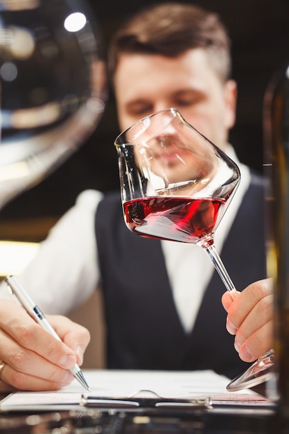 Un hombre maduro con chaleco llena un formulario en papel sosteniendo una elegante copa de delicioso vino tinto natural en la mesa en el primer plano del restaurante