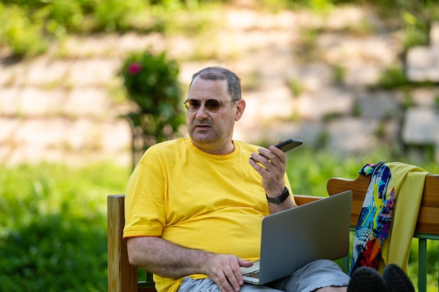 Hombre maduro con computadora portátil y teléfono inteligente trabajando afuera en el concepto de oficina en casa verde jardín