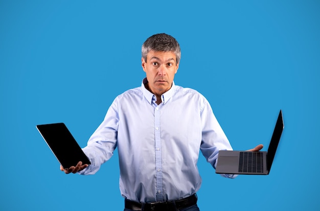 Hombre maduro desconcertado que sostiene la tableta y la computadora portátil que elige el tiro del estudio