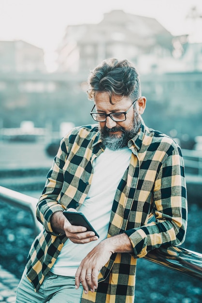 Hombre maduro escribe y lee en el teléfono móvil de pie al aire libre Tecnología y concepto de estilo de vida de la gente moderna Estilo hipster con barba masculina y anteojos usa la aplicación en el celular y envía mensajes de chat