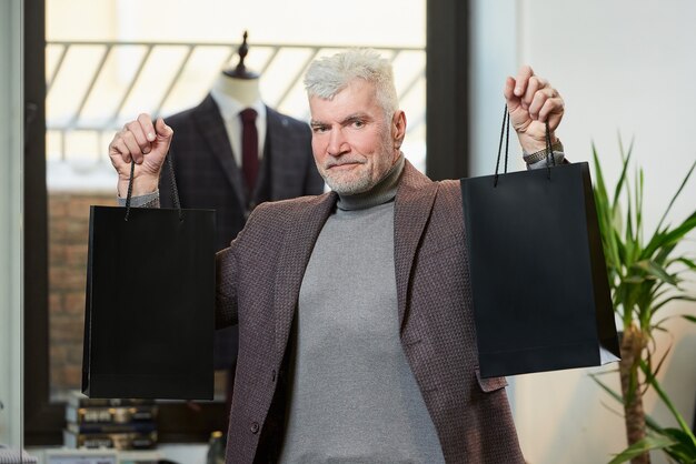 Un hombre maduro feliz con cabello gris y físico deportivo muestra dos bolsas de papel negras con compras en una tienda de ropa. Un cliente masculino con barba viste un traje de lana en una boutique