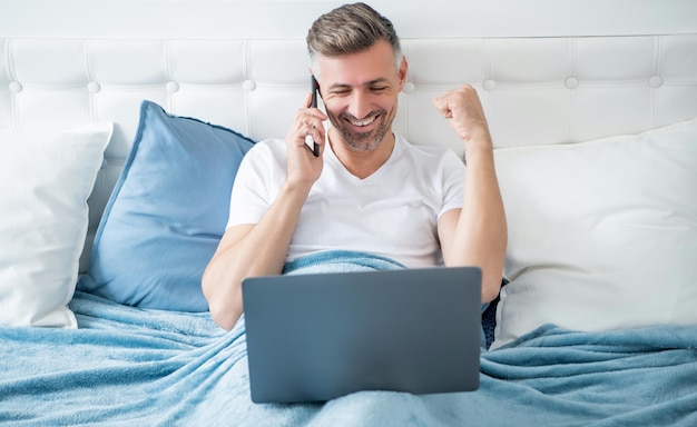 Un hombre maduro feliz hablando por teléfono y trabajando en una laptop en la cama