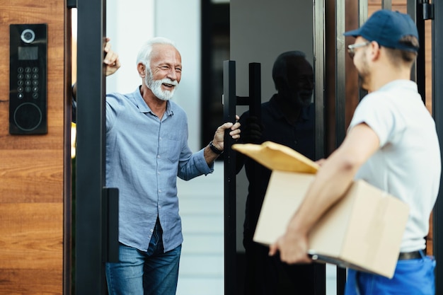 Hombre maduro feliz parado en la puerta y hablando con el mensajero que le está entregando un paquete