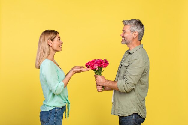 Hombre maduro feliz presentando flores a su esposa para el Día de San Valentín o aniversario sobre fondo de estudio amarillo