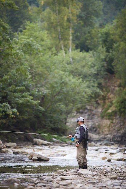 Hombre maduro con gorra y uniforme con varilla para pescar al aire libre. Pesca de verano en río rápido. La naturaleza verde de la montaña alrededor.