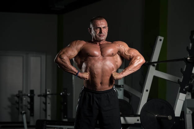 Hombre maduro guapo de pie fuerte en el gimnasio y flexionando los músculos Muscular culturista atlético Fitness masculino posando después de los ejercicios