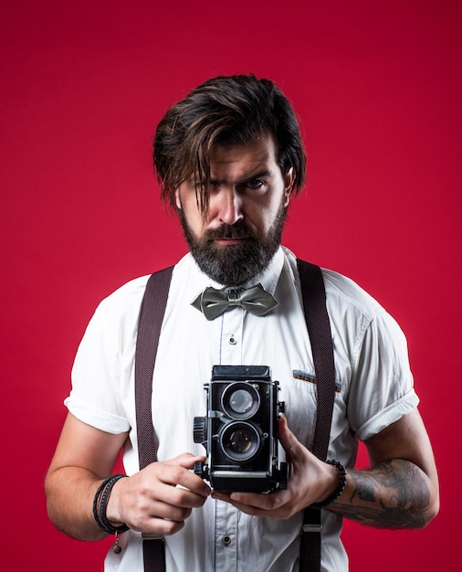 un hombre maduro y guapo con tirantes sosteniendo una cámara retro mientras se enfrenta a un fondo rojo, fotografiando.