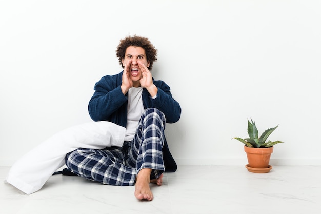 Hombre maduro en pijama sentado en el piso de la casa gritando emocionado al frente.