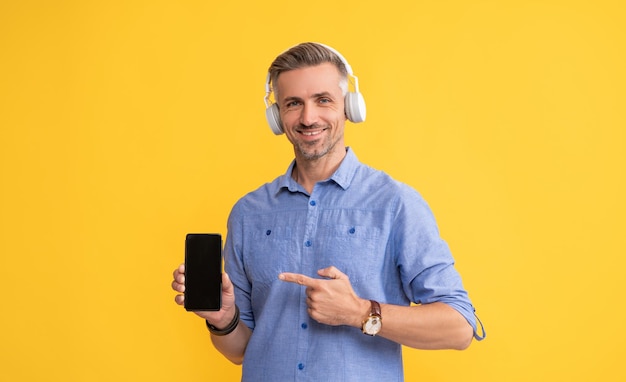 Hombre maduro sonriente escucha música en auriculares señalando con el dedo en la nueva aplicación de teléfono inteligente