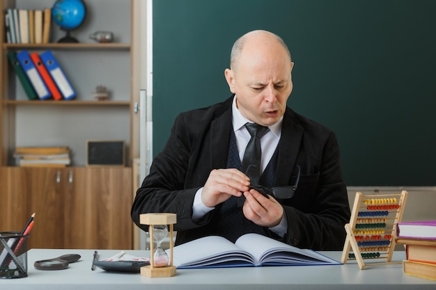 Hombre maestro revisando el registro de clase limpiando sus anteojos luciendo confiado sentado en el escritorio de la escuela frente a la pizarra en el aula