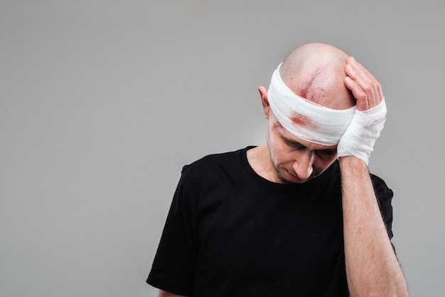 Un hombre maltratado con una camiseta negra está de pie contra una pared gris, sosteniendo su cabeza dolorida con las manos envueltas alrededor.
