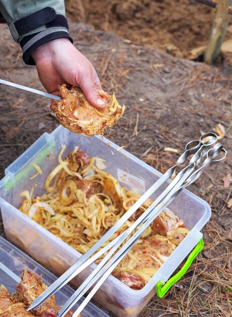 Foto hombre con una mano sosteniendo la carne, con la otra - ensartando brocheta. enhebrar carne en una brocheta en la naturaleza. cámping