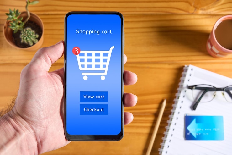 Foto hombre con la mano sosteniendo un teléfono móvil con interfaz de tienda en línea en la pantalla icono del carrito de compras y botones para ver el carrito y la caja tarjeta de crédito y accesorios en la mesa en el fondo vista superior