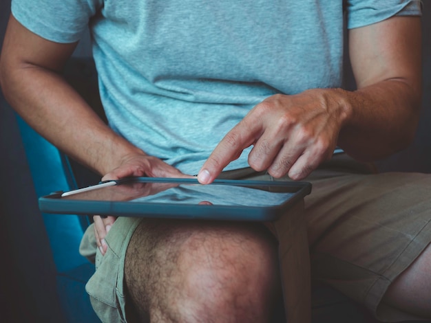 Hombre de mano con tableta digital. Una persona joven, un hombre con camisa gris casual y pantalones cortos sentado e interactuando con la tecnología en casa. Hombre de negocios de relajación trabajando en línea en cualquier lugar concepto.