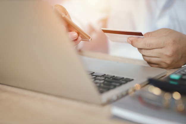 Hombre manos en camisa blanca sentado y sosteniendo la tarjeta de crédito y usando la computadora portátil en la mesa para pago en línea o compras en línea. Hombre de negocios mediante teléfono móvil mientras está sentado. Concepto de banca electrónica