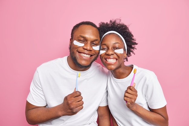 El hombre mantenga cepillos de dientes aplique parches de belleza debajo de los ojos vestidos con camisetas básicas blancas casuales aisladas en rosa