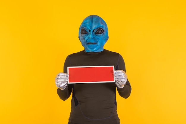 Hombre con máscara alienígena con cabeza azul sosteniendo un cartel rojo Concepto de extraño extraterrestre divertido informativo extraño y raro