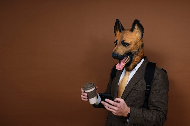 Un hombre con una máscara de cabeza de perro de látex y una mochila manejando un teléfono inteligente, llevando una taza de café en la mano