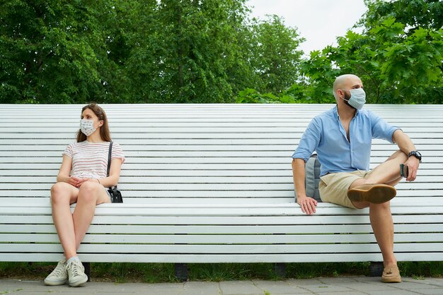 Un hombre con una máscara facial está sentado en un banco manteniendo una distancia social con una mujer