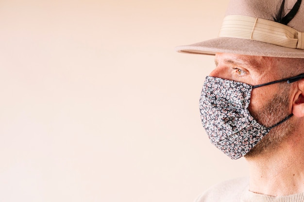 Hombre con máscara de flores y sombrero contra la pared beige durante el coronavirus